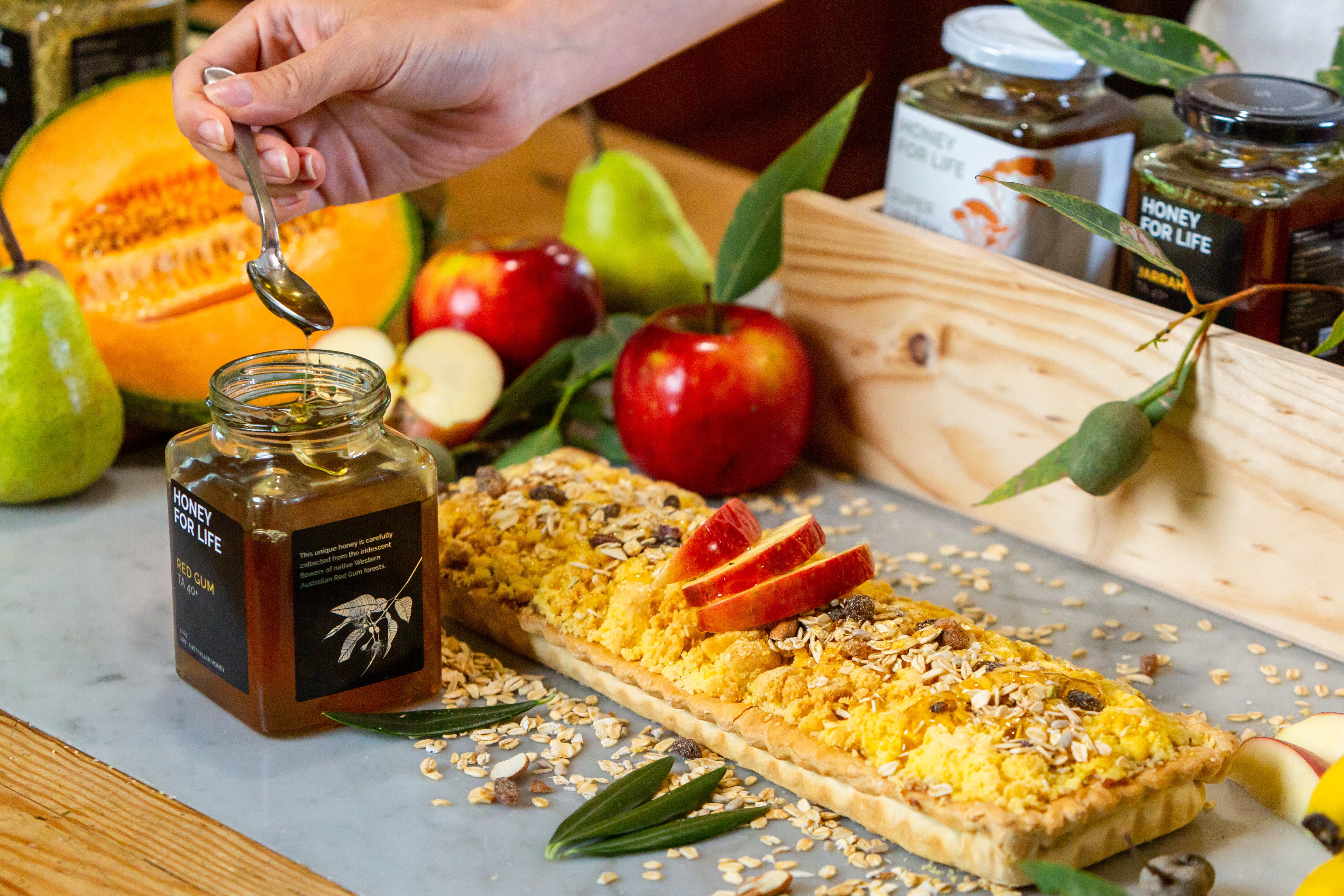 Bord med frugter og tærte, et glas med Honey For Life der hældes over tærten med en ske